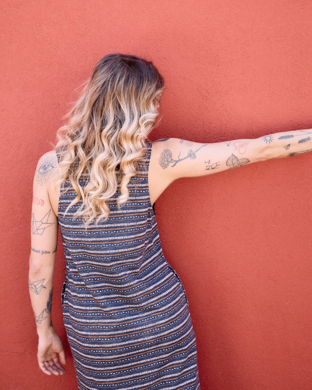 A person with long, wavy hair, wearing a sleeveless striped dress featuring a tribal stripe pattern, stands facing a red wall with their right arm extended to the side, showing multiple tattoos on their arms and back. The dress is the Saltrock Tribal Stripe - Womens Midi Dress - Grey.