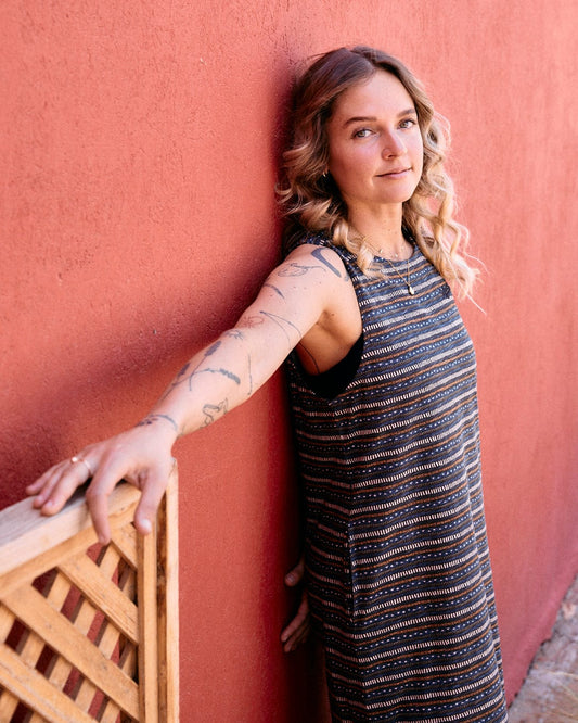 A woman with wavy hair leans against a red wall, extending her arm over a wooden lattice. She is wearing the Tribal Stripe - Womens Midi Dress - Grey from Saltrock in 100% cotton and has visible tattoos on her arms.