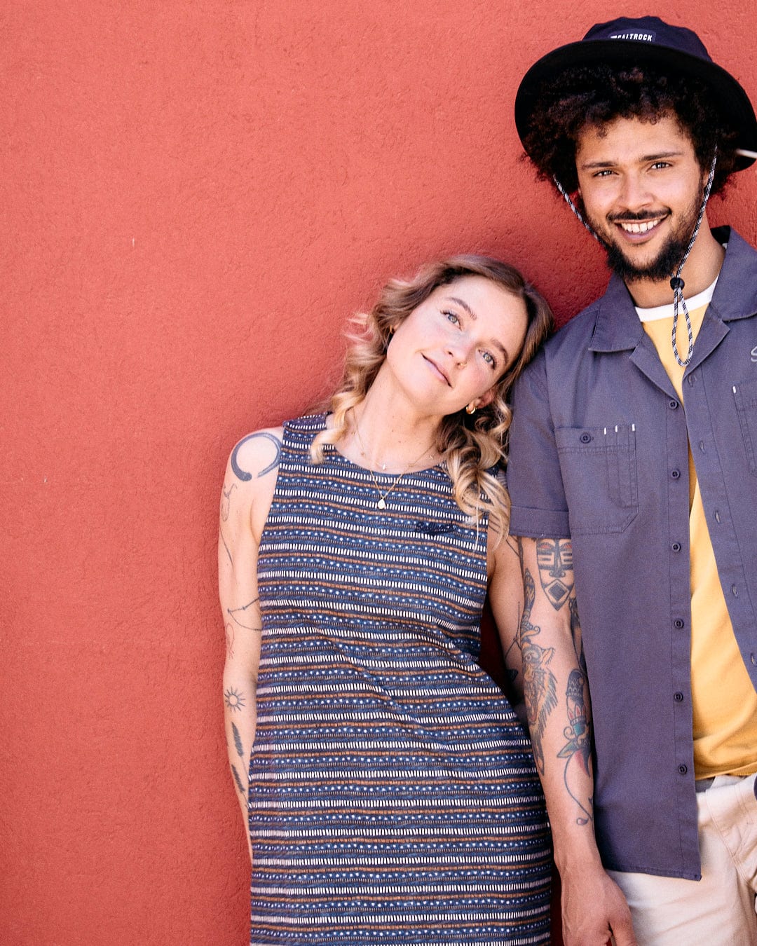 A woman in a Saltrock Tribal Stripe - Womens Midi Dress - Grey and a man in a blue shirt and hat stand next to a red wall. The man, with Saltrock branded embroidery on his shirt, is smiling while the woman has a neutral expression. Both have visible tattoos.