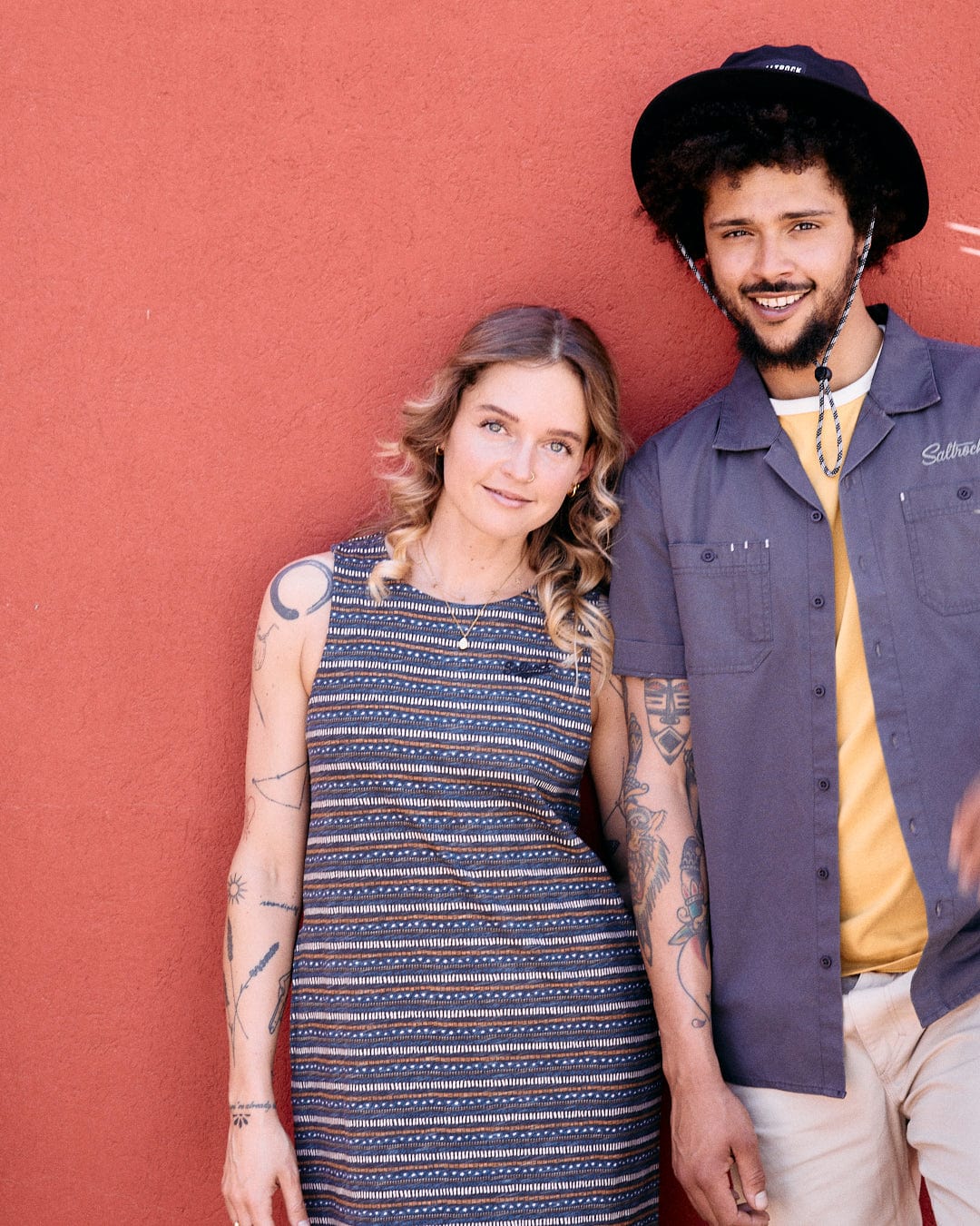 A woman in a Saltrock Tribal Stripe - Womens Midi Dress - Grey and a man in a hat and gray shirt with tattoos stand in front of a red wall, smiling.