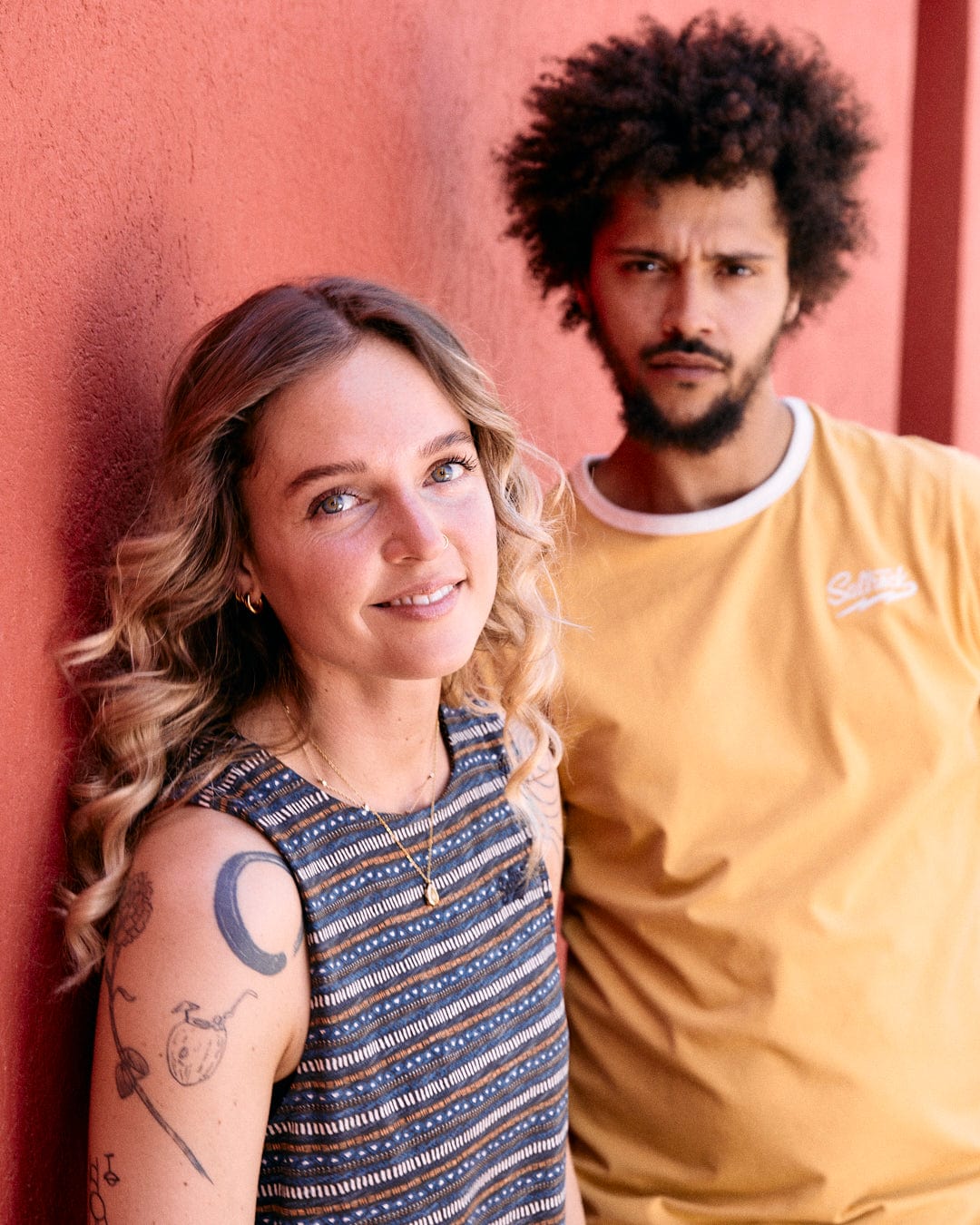 Two people are standing against a red wall. The woman is smiling, has wavy blonde hair, and wears the Saltrock Tribal Stripe - Womens Midi Dress - Grey. The man, with curly hair and a beard, wears a mustard yellow shirt made of 100% cotton and looks at the camera.