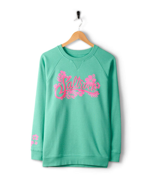 A green sweatshirt made of 100% cotton, featuring Saltrock branding and a pink floral design printed on the front, hangs on a wooden hanger against a white background. The Tropic - Womens Sweat - Green by Saltrock is machine washable for easy care.