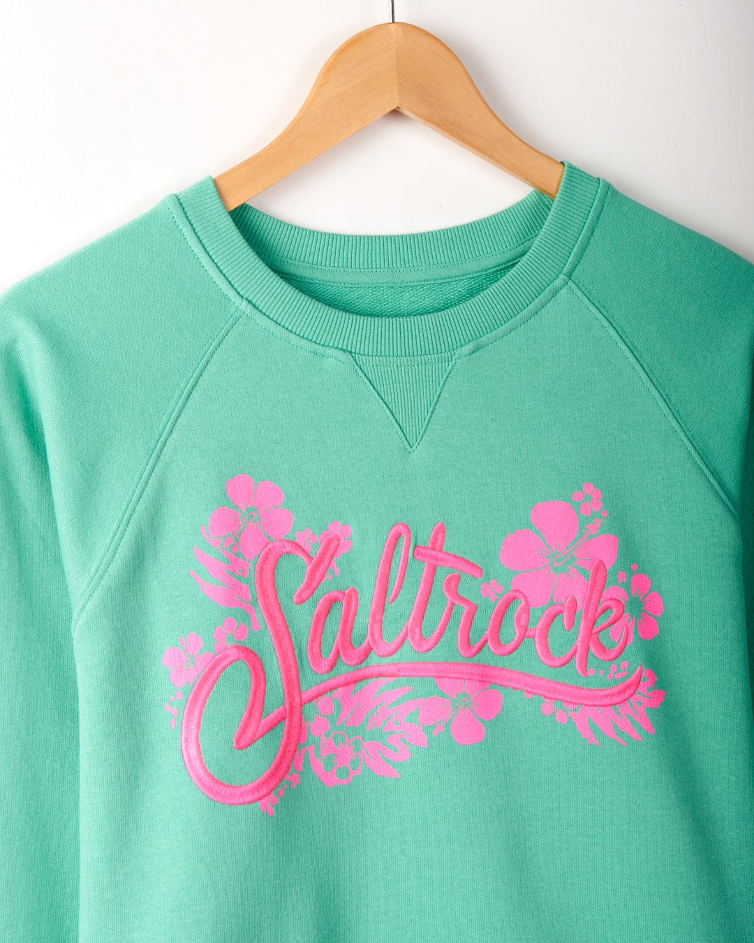 A Tropic - Womens Sweat - Green on a wooden hanger featuring the word "Saltrock" in pink cursive with floral accents, made from 100% cotton and machine washable.