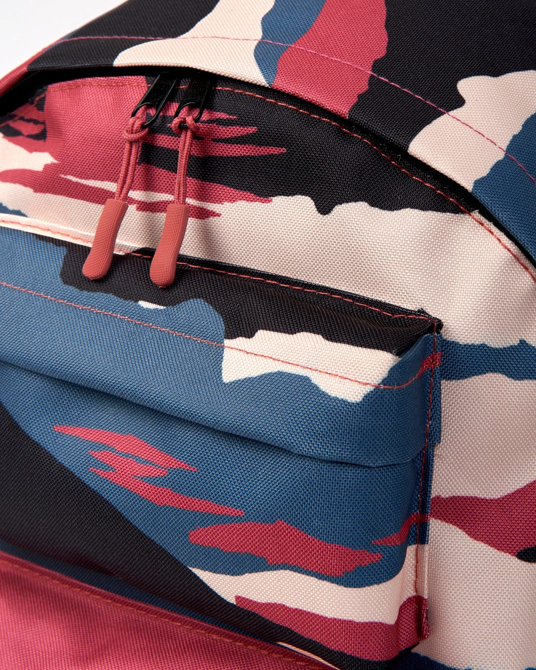 Close-up of the **Uni Backpack by Saltrock** featuring a vibrant camouflage pattern in shades of blue, pink, peach, and black. The image highlights the top zippered pocket and dual zipper pullers. This backpack includes a water-resistant laptop sleeve for added functionality.