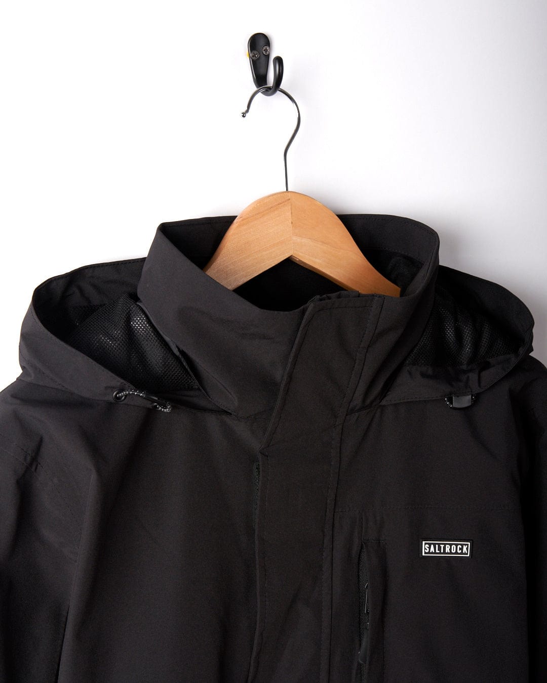 The Whistler II Men's Waterproof Hooded Jacket by Saltrock, displayed in black on a hanger against a white wall, features a convenient packable hood. This jacket is both waterproof and breathable, making it ideal for staying comfortable and dry in unpredictable weather.