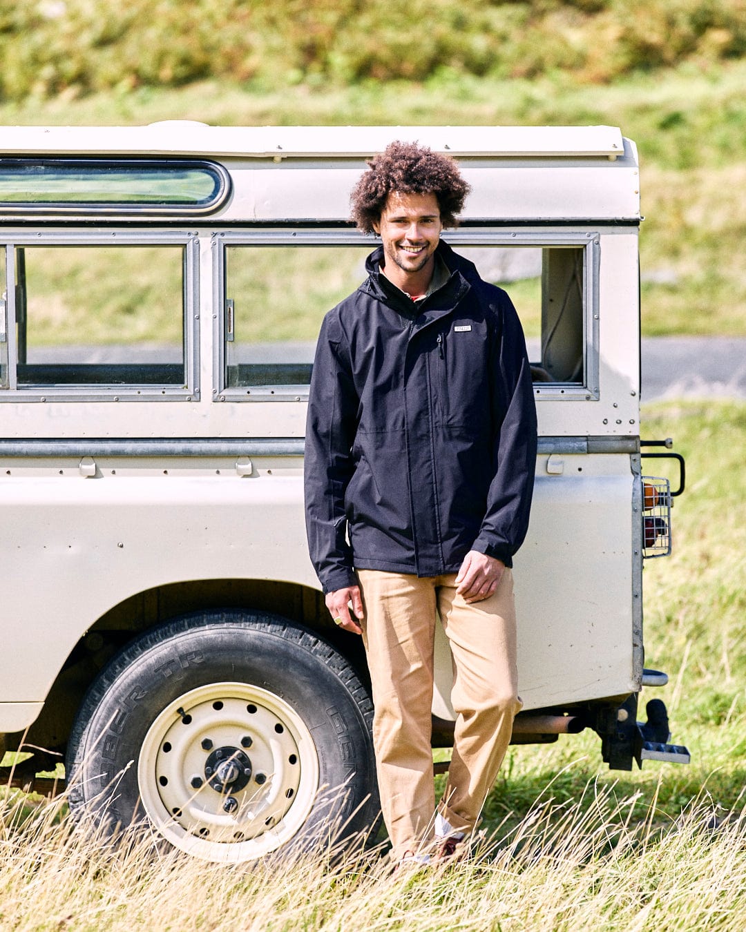 A man wearing the Saltrock Whistler II Men's Waterproof Hooded Jacket in black and beige pants stands smiling beside a vintage white off-road vehicle in a grassy area.