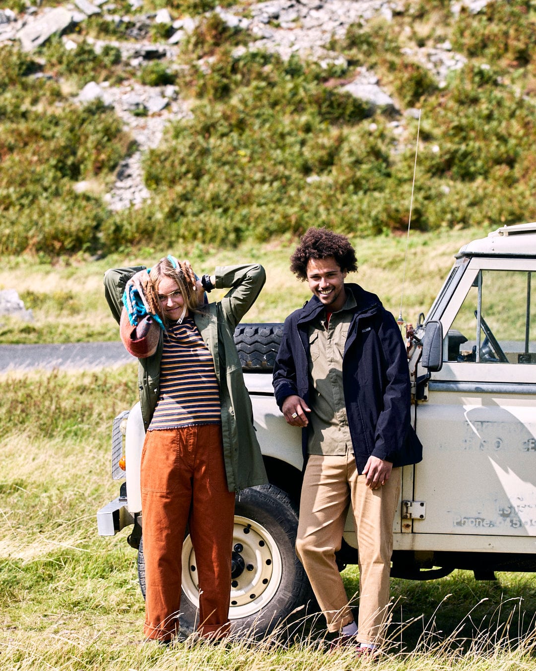 Two people stand beside an off-road vehicle in a grassy area. One adjusts their scarf, while the other smiles with hands in pockets, wearing the Saltrock Whistler II Mens Waterproof Hooded Jacket in Black. A rugged landscape serves as the backdrop.