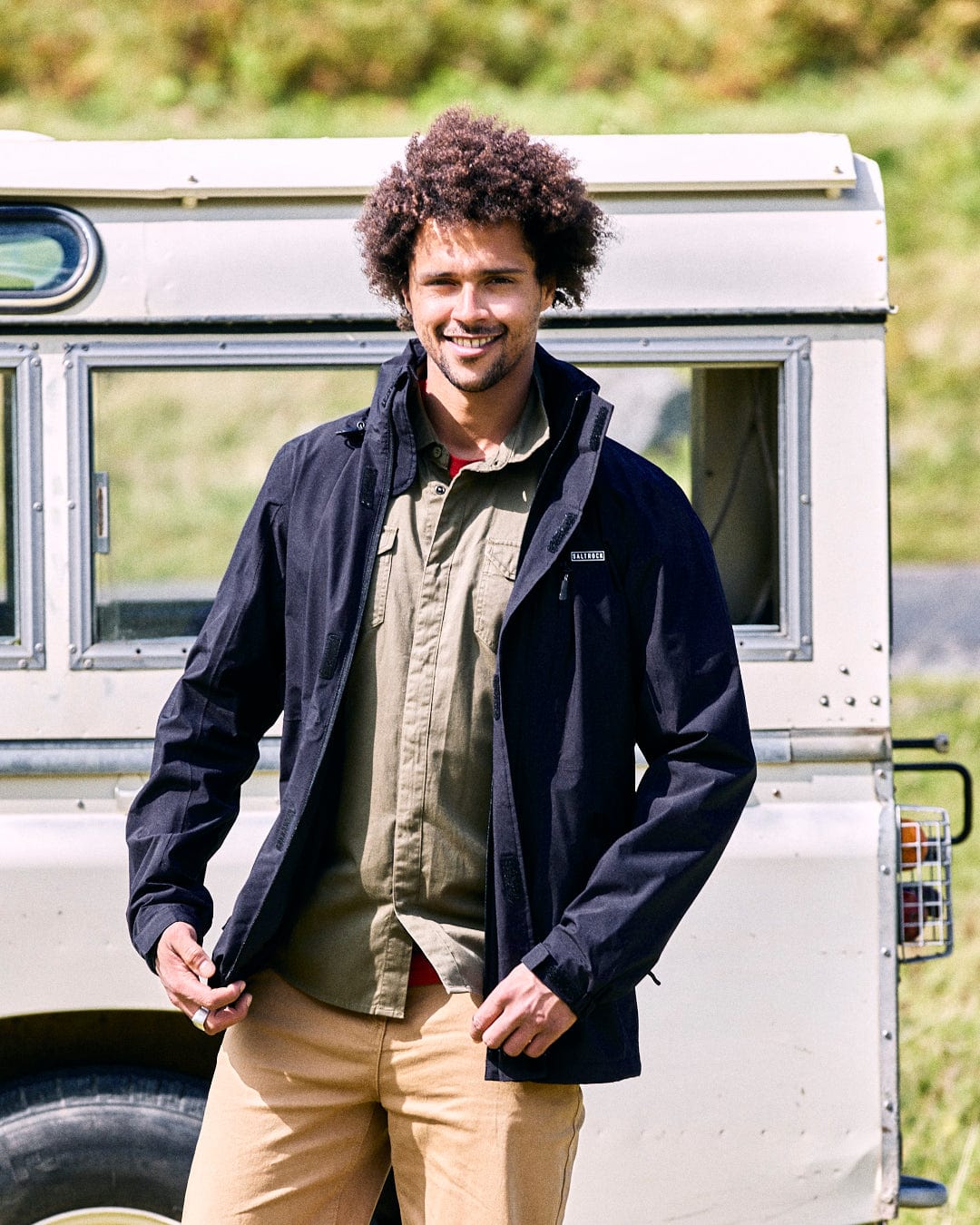 A man with curly hair is wearing the Whistler II men's black waterproof hooded jacket by Saltrock and khaki pants, smiling as he stands in front of a white vehicle.