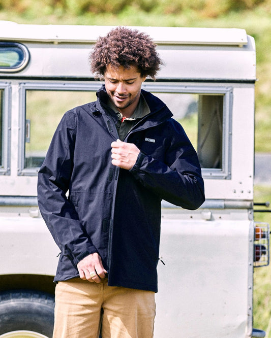 A person with curly hair zips up a sleek, breathable Whistler II men's waterproof hooded jacket in black by Saltrock, while standing in front of a vintage vehicle.