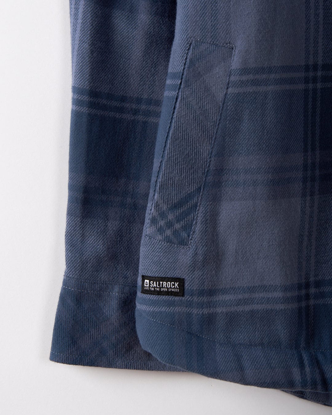 A close-up of the Woody men's Sherpa-lined shacket in mid blue showcases a blue check plaid fabric with a visible pocket, highlighted by the small "Saltrock" label near the hem. Crafted from recycled materials, this shacket effortlessly combines style and sustainability.