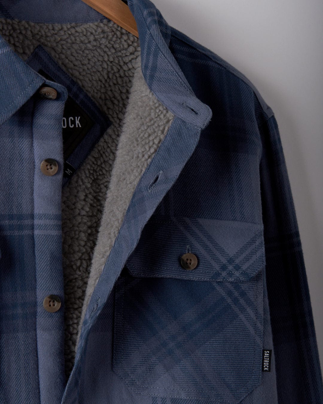 Close-up of the Saltrock "Woody - Men's Sherpa Lined Shacket - Mid Blue" hanging on a wooden hanger. The shacket boasts a blue check plaid pattern made with recycled materials and includes cozy sherpa lining, along with a front pocket and button details.