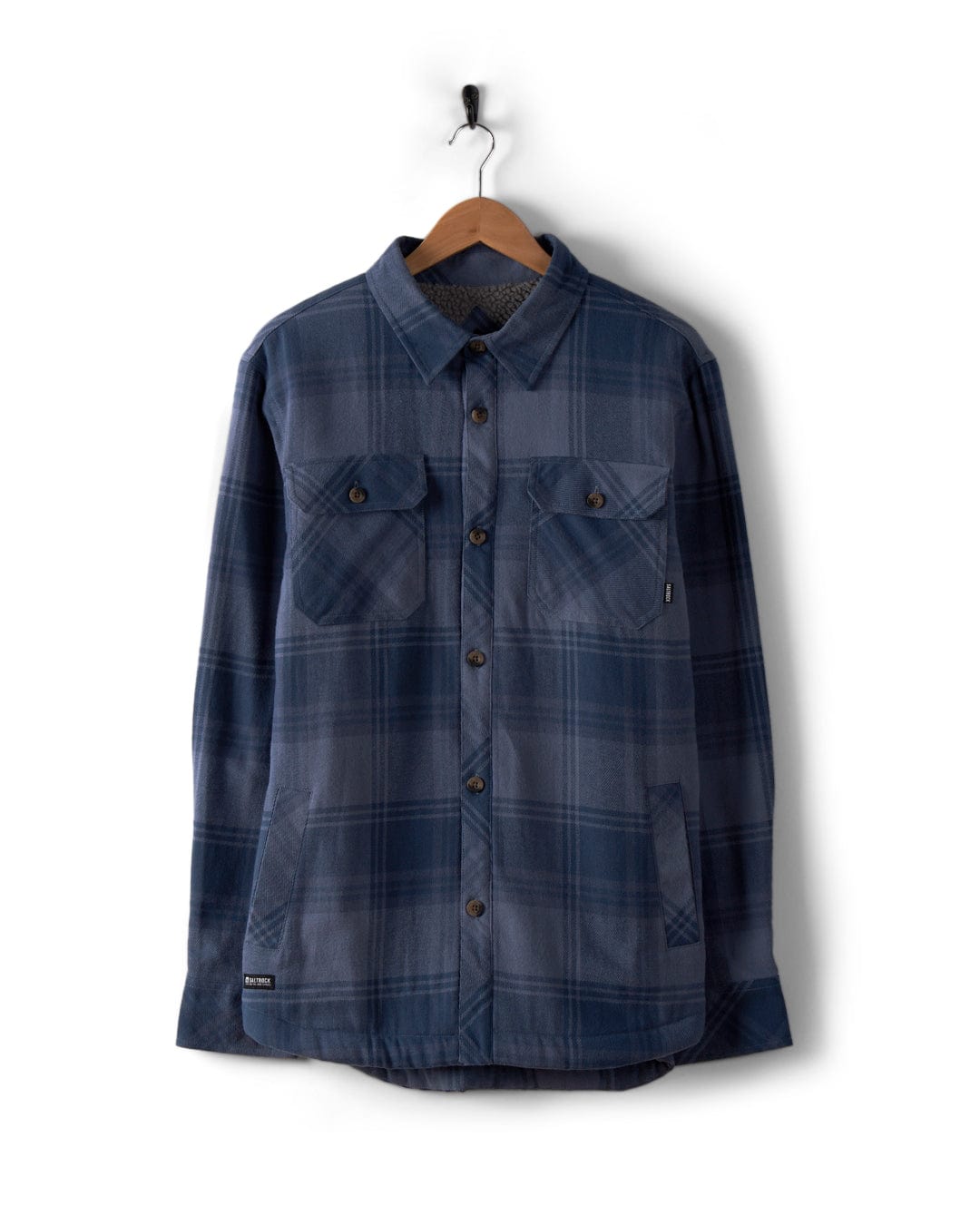 The "Woody" men's Sherpa-lined shacket in mid-blue from Saltrock hangs on a wooden hanger against a white background.