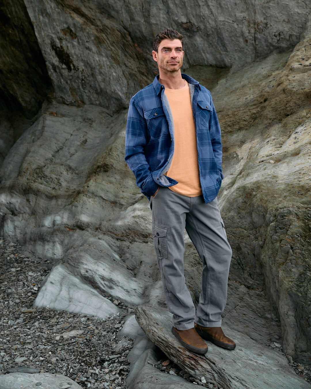 A man stands on a rugged surface, showcasing the Saltrock "Woody" Sherpa Lined Shacket in Mid Blue over a beige shirt, paired with gray pants and brown boots.