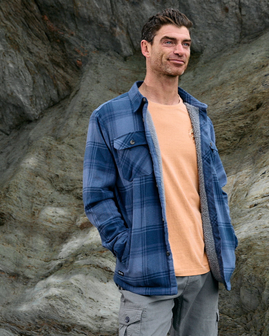 A person wearing the Saltrock Woody men's sherpa-lined shacket in mid blue and a beige shirt stands in front of large rocks.