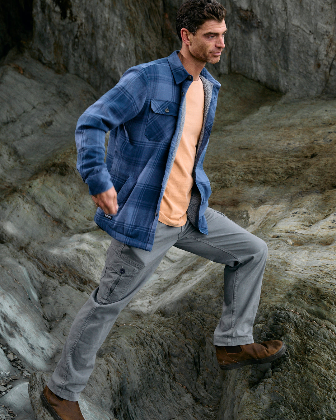 A man wearing the Woody shacket by Saltrock, featuring a blue check plaid design and made with sherpa lining crafted from recycled materials, pairs it with gray pants as he steps on rocks outdoors.