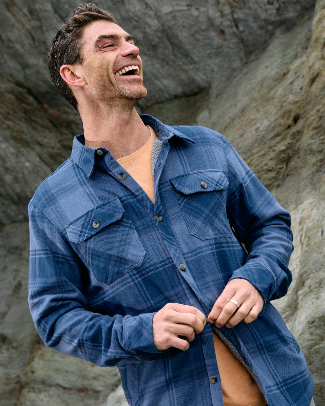 A man confidently enjoys the outdoors while wearing the Saltrock Woody - Mens Sherpa Lined Shacket in Mid Blue and a vibrant orange t-shirt. He stands against a backdrop of rock formations, smiling as he holds the bottom button of his shacket crafted from recycled materials.