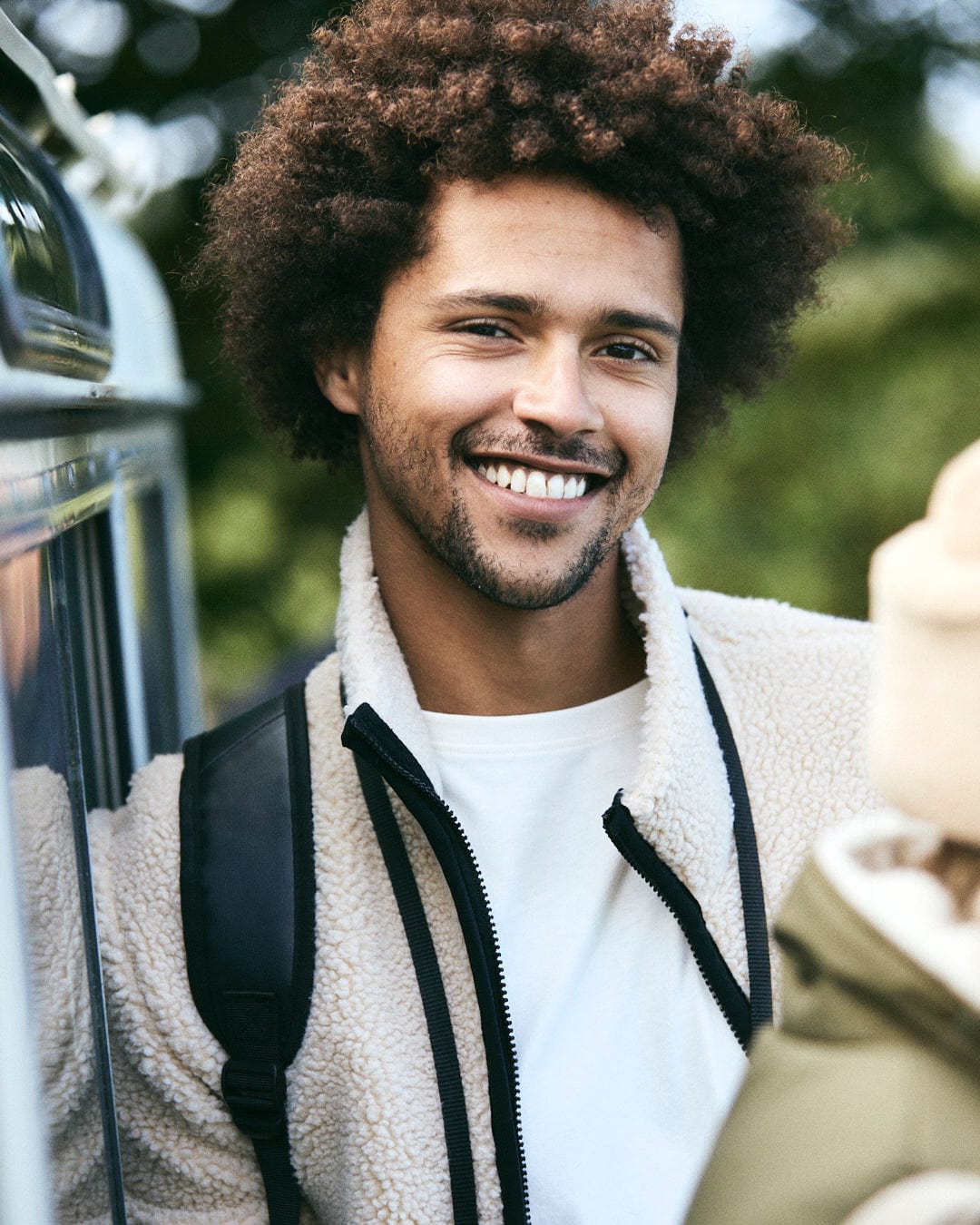 A person with curly hair stands outdoors, smiling and wearing the cozy "Wye 2 - Mens Recycled Fleece - Cream" by Saltrock, effortlessly carrying a backpack.