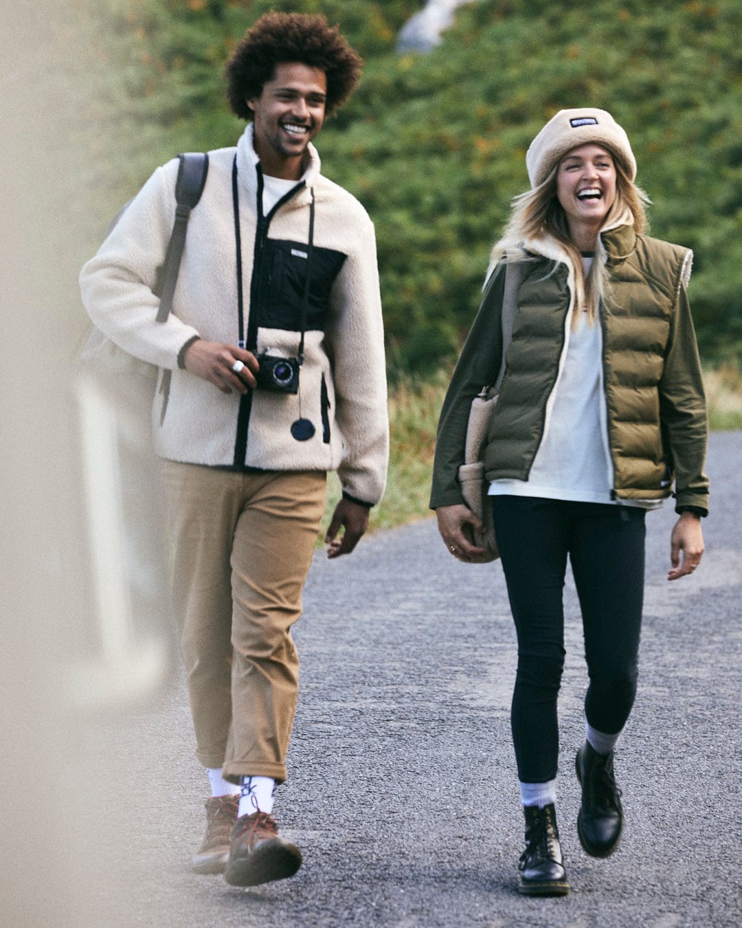 A man and woman stroll joyfully, with the man donning a Saltrock Wye 2 Men's Recycled Fleece in cream, camera in hand, and the woman matching his look with a green vest and wool hat. They stylishly enjoy the crisp outdoors in recycled materials.