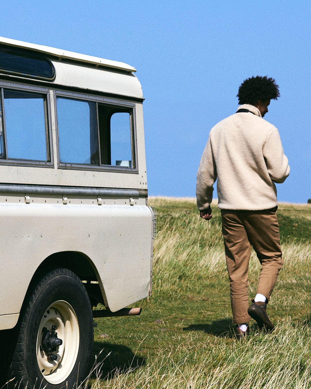 A person wearing the Saltrock Wye 2 Men's Recycled Fleece in cream walks away from a vintage beige vehicle parked on the grass under the clear blue sky.