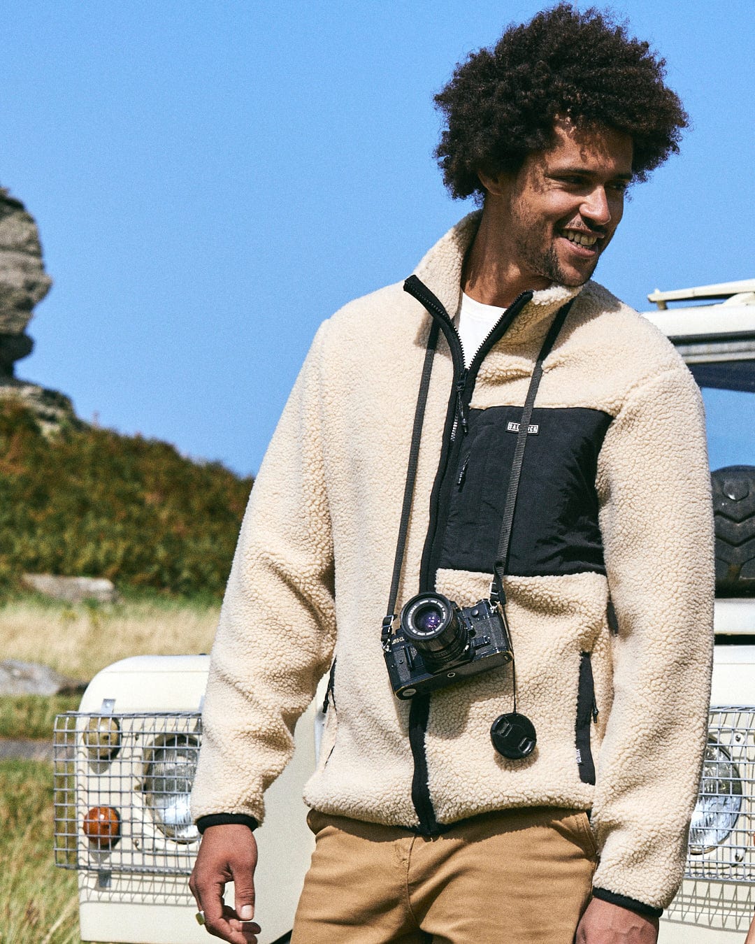 A man in a Saltrock Wye 2 Men's Recycled Fleece - Cream stands before an off-road vehicle, vintage camera in hand, with grassy plains and striking rock formations under a clear blue sky, his eco-friendly outerwear perfectly complementing the rugged landscape.