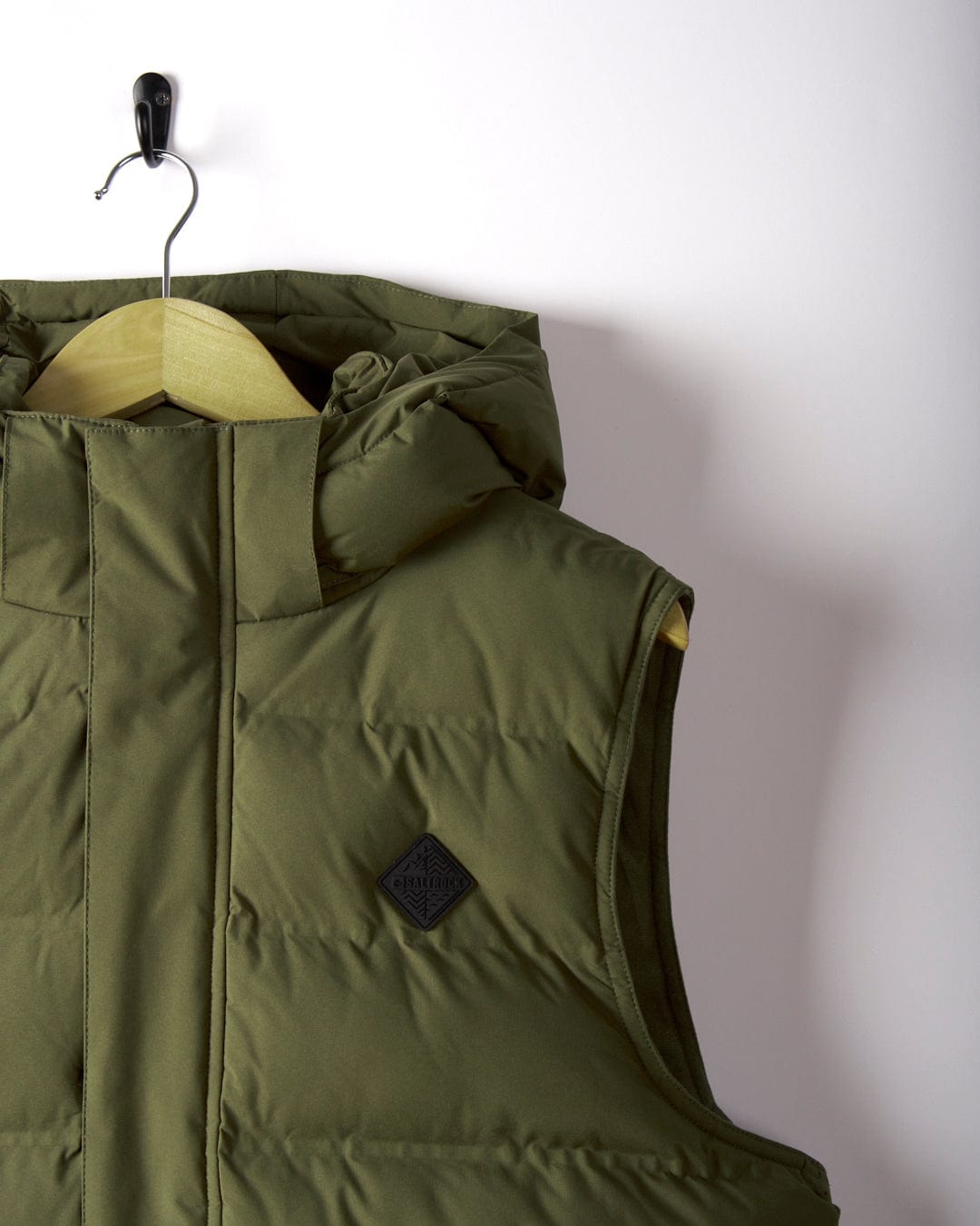 The Xavier - Mens Padded Gilet by Saltrock, in olive green, hangs on a wooden hanger against a white wall. Made from water-resistant polyester, it includes a detachable hood and displays a small black logo patch on the chest.