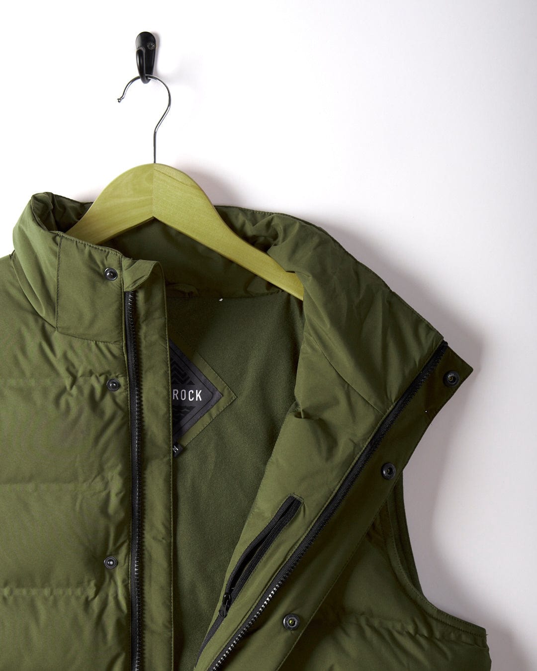 The Xavier Men's Padded Gilet in green by Saltrock, featuring a detachable hood and made from water-resistant polyester, is elegantly displayed on a wooden hanger from a black wall hook.