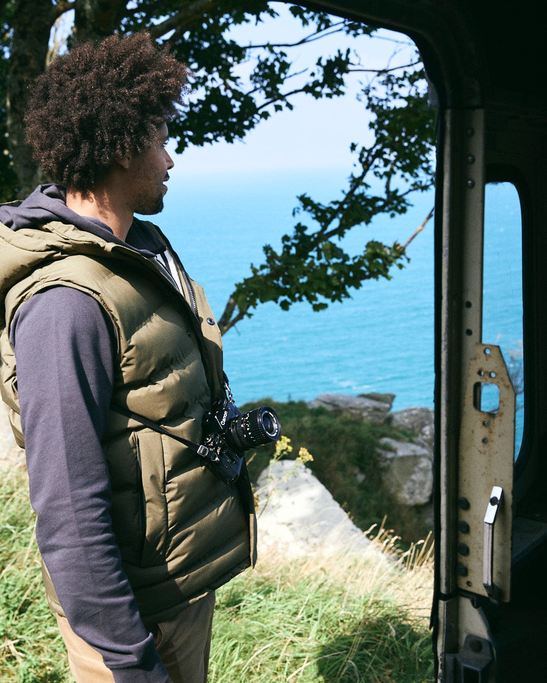 A person wearing the Saltrock Xavier Men's Padded Gilet in green, with a camera, stands inside a doorway, gazing out at the ocean and trees.