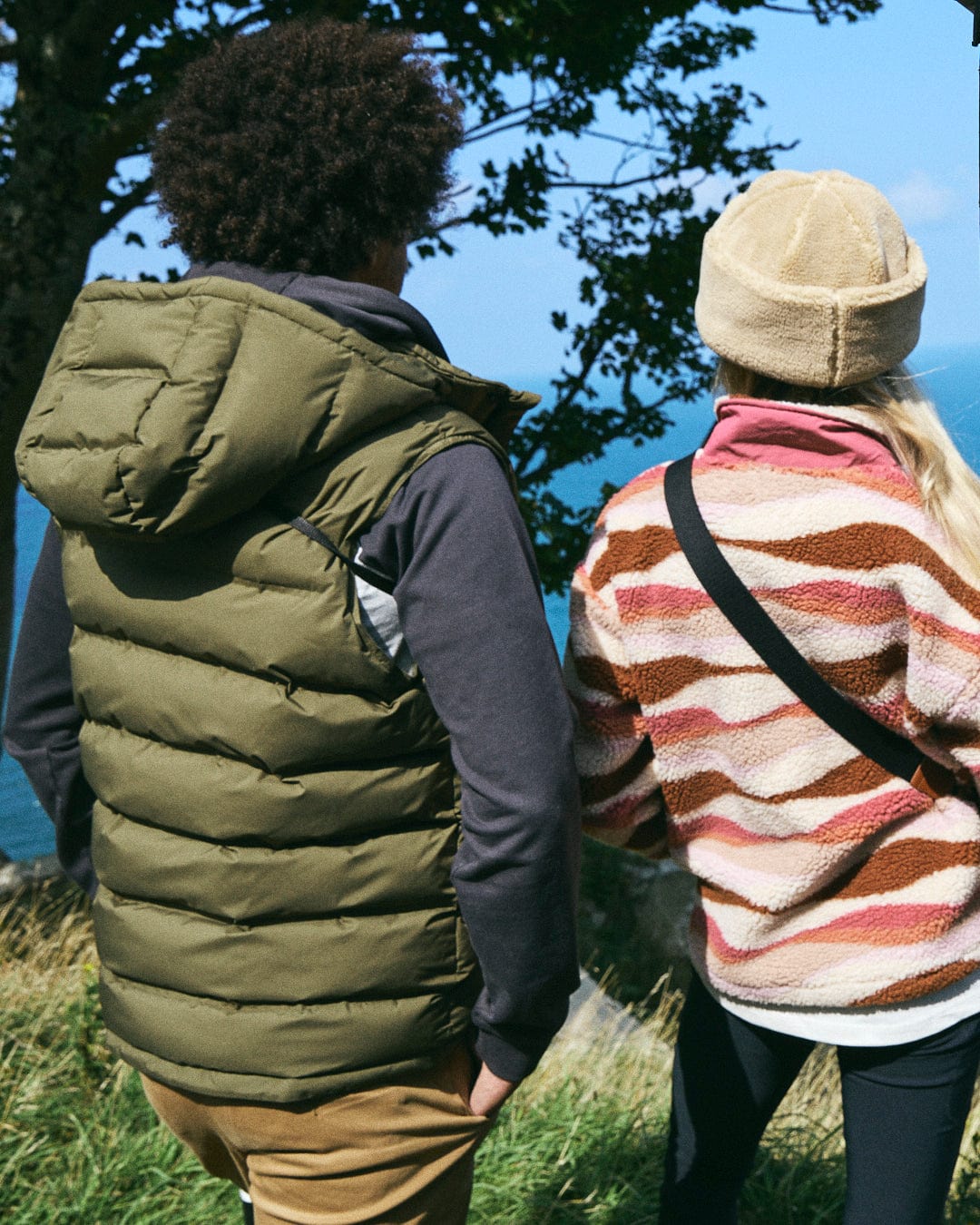 Two individuals are wearing Saltrock's green Xavier Men's Padded Gilet, which features a polyester construction and detachable hood, as they stand on a grassy hill, taking in the scenic ocean view.