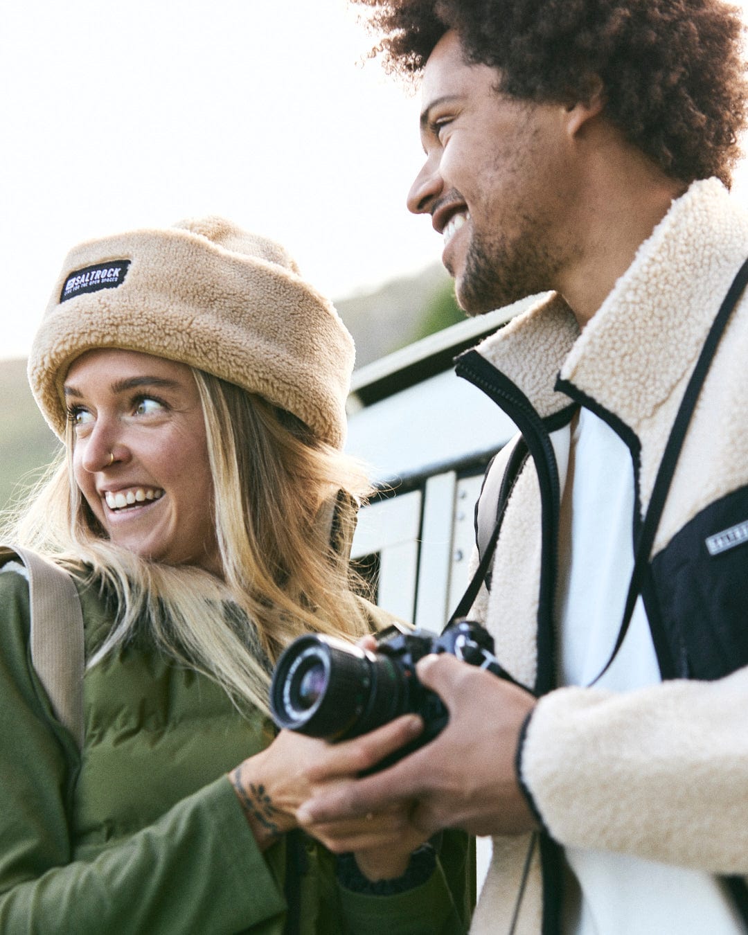 Two people, dressed in Saltrock's cozy Wye 2 Men's Recycled Fleece in cream, enjoy the outdoors on a brisk day. One holds a camera as they both smile, their warm, machine washable sherpa fleece keeping them snug.