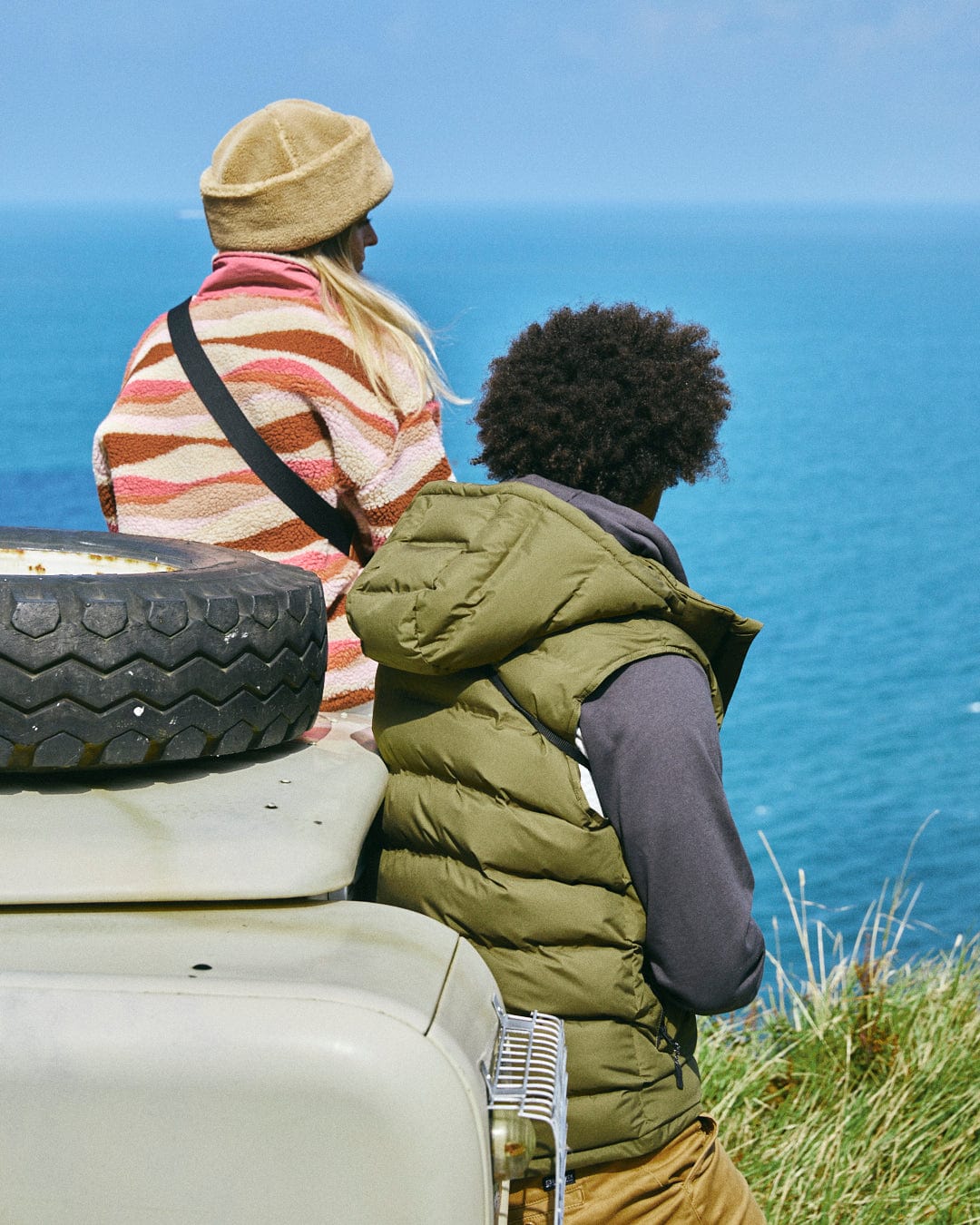 Two people sit on a vehicle by the coast, gazing at the ocean. They're comfortably dressed in casual, warm clothing from Saltrock, featuring water-resistant polyester, and they relax next to a spare tire, enjoying the serene view. One of them is wearing the Xavier Men's Padded Gilet in green.