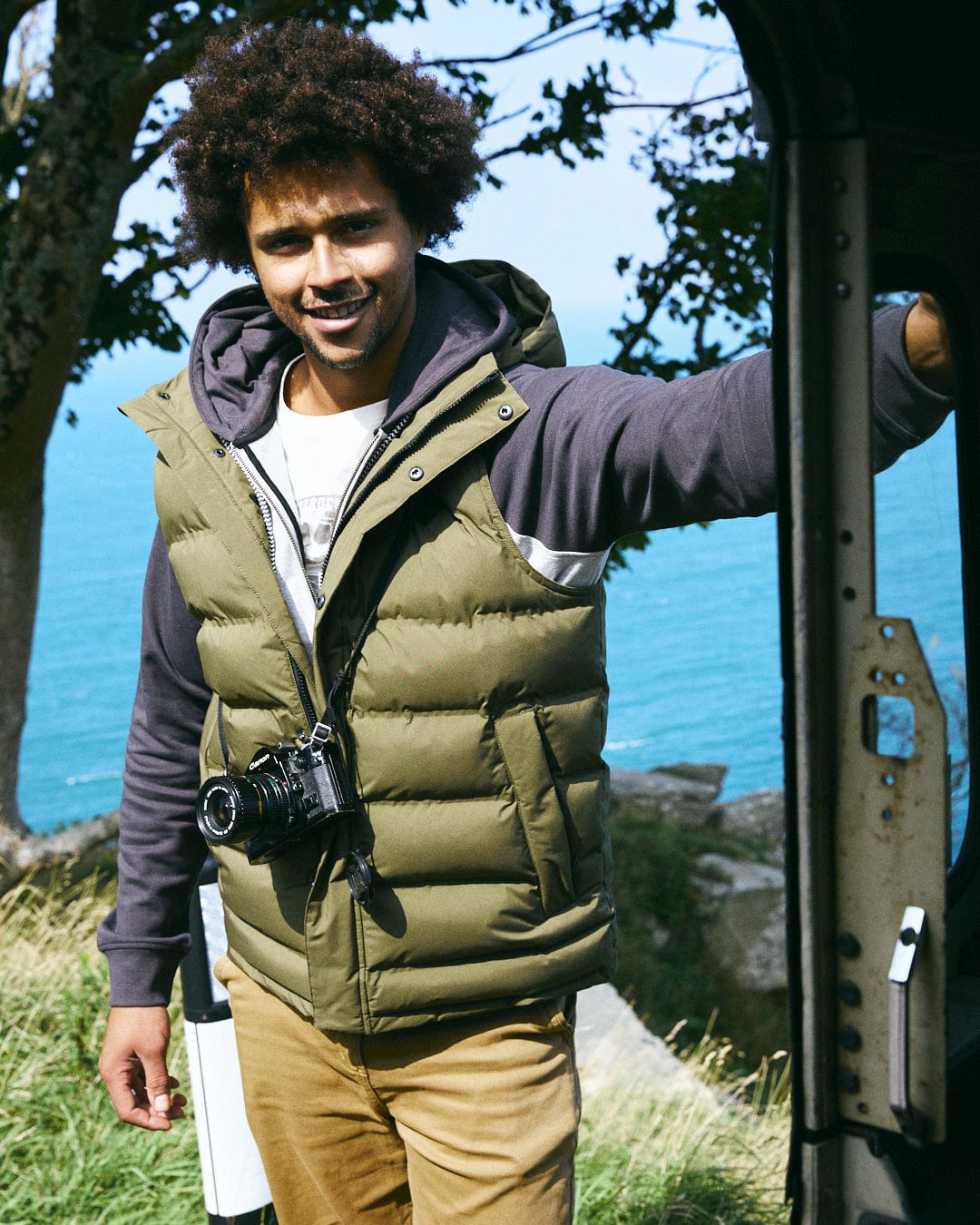 A person wearing the Saltrock Xavier Men's Padded Gilet in green, which features a detachable hood, holds a camera while standing by a door with the ocean and trees in the background.