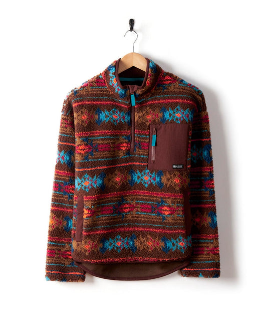 Hanging on a hook, the Zella Aztec by Saltrock is a cosy recycled fleece sweater for women, showcasing a vibrant Aztec print in shades of red, blue, and brown.