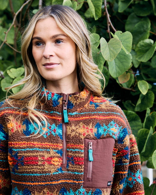 Wearing the Zella Aztec recycled fleece by Saltrock, a woman stands amidst green leaves, gazing to the side with intrigue.