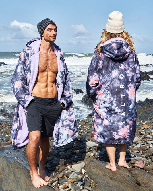 On a rocky beach, two people sport vibrant Saltrock Recycled Changing Robe in Pink Tie Dye. The shirtless man pairs his robe with black shorts, while the woman faces away from the camera. They both wear beanies as they enjoy the coastal view.