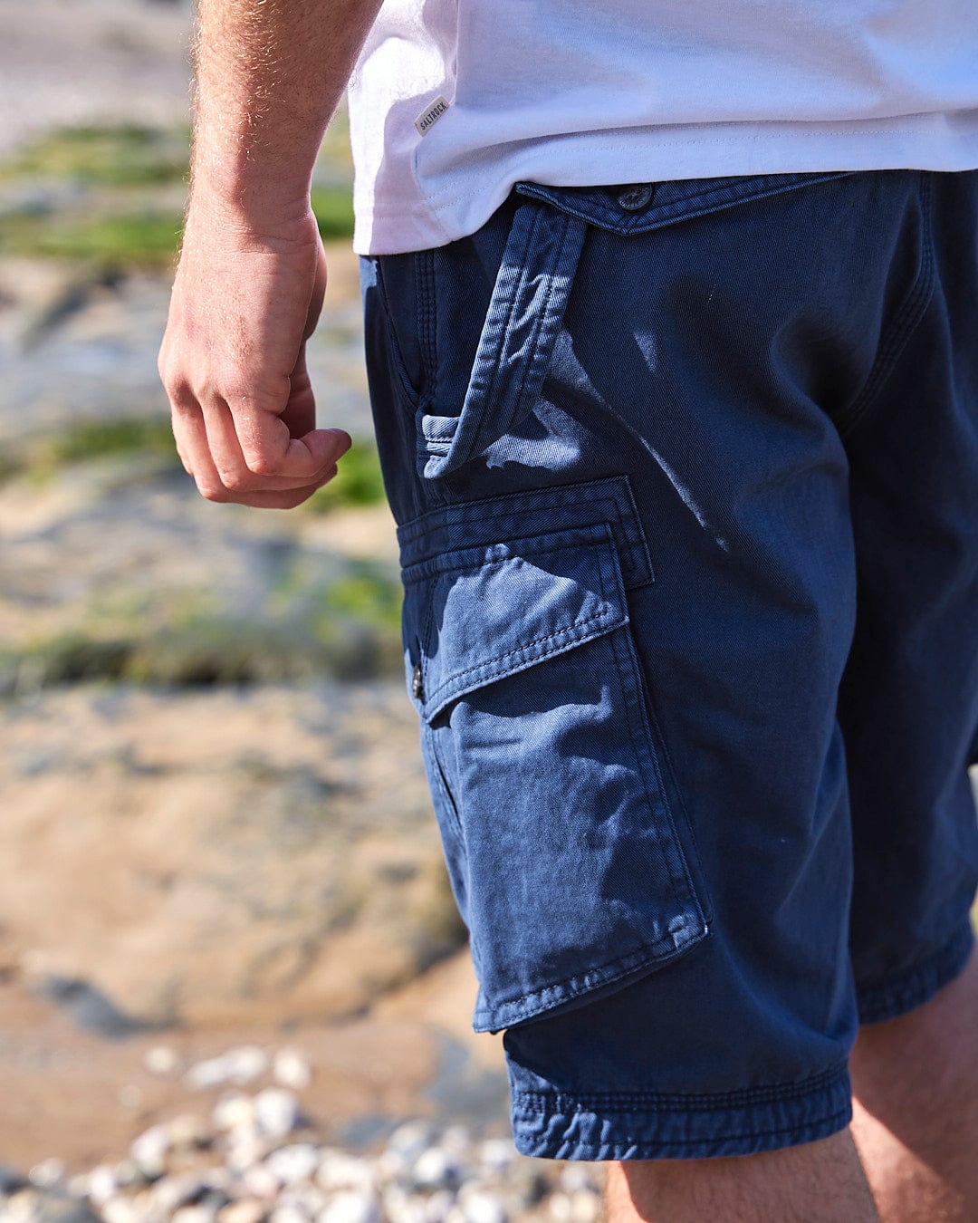 A man is standing on a beach in practical style, wearing Penwith II - Mens Cargo Shorts - Blue with a Saltrock twist.